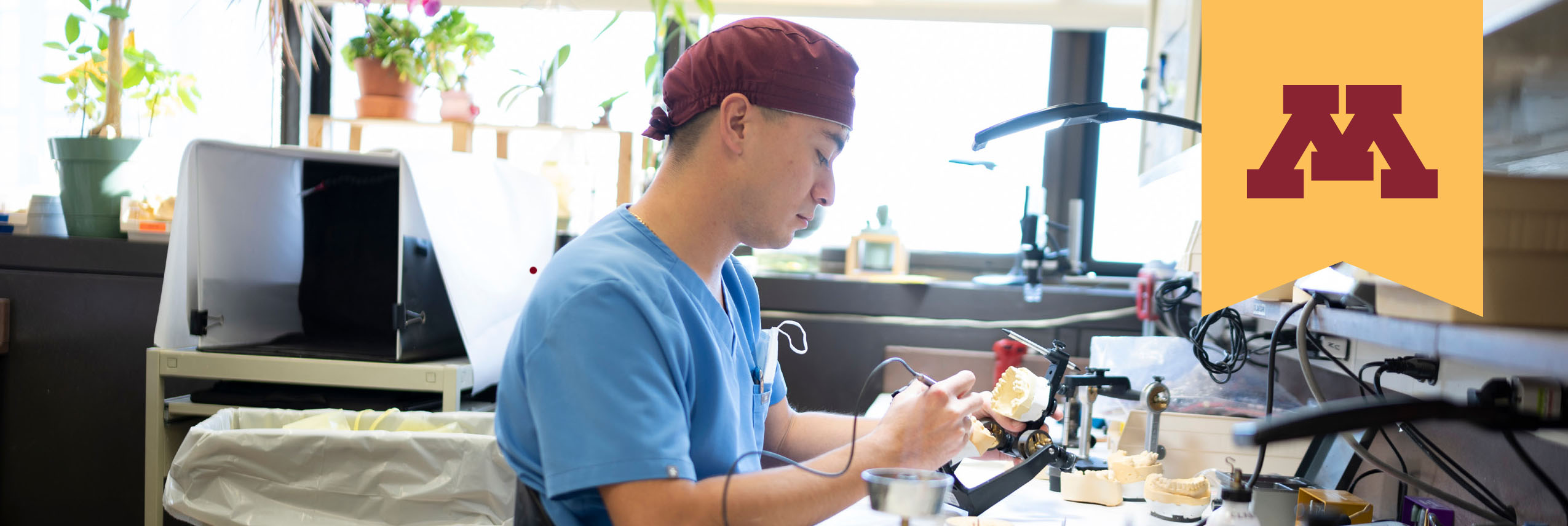 Gold banner with UMN maroon block M placed next to dental student in blue scrubs who shapes a dental impression