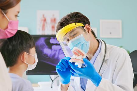 A dentist shows a model of teeth to a young child and his mother.