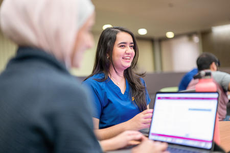 A student smiles and talks with other students while another student works on her computer, blurred in the foreground