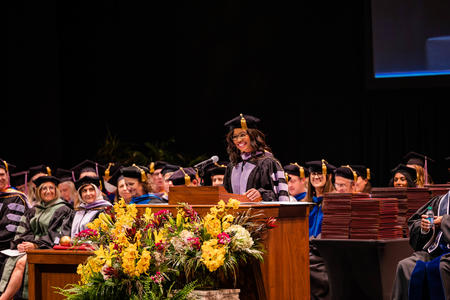 Monica Anderson speaks behind a podium in regalia