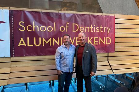 Ryan anderson and Mark Bachman in front of an Alumni Weekend banner