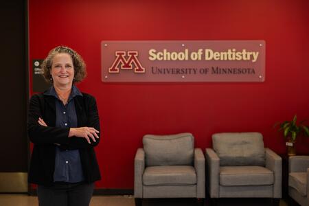 Kristin Artinger stands and smiles in front of a School of Dentistry sign on a maroon wall