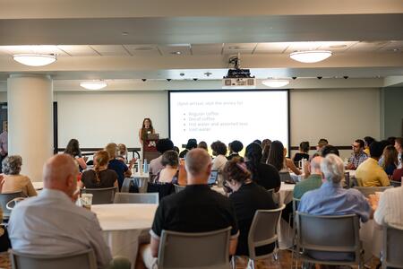 A full room of attendees listen to Sarah Aitken as she presents from a podium