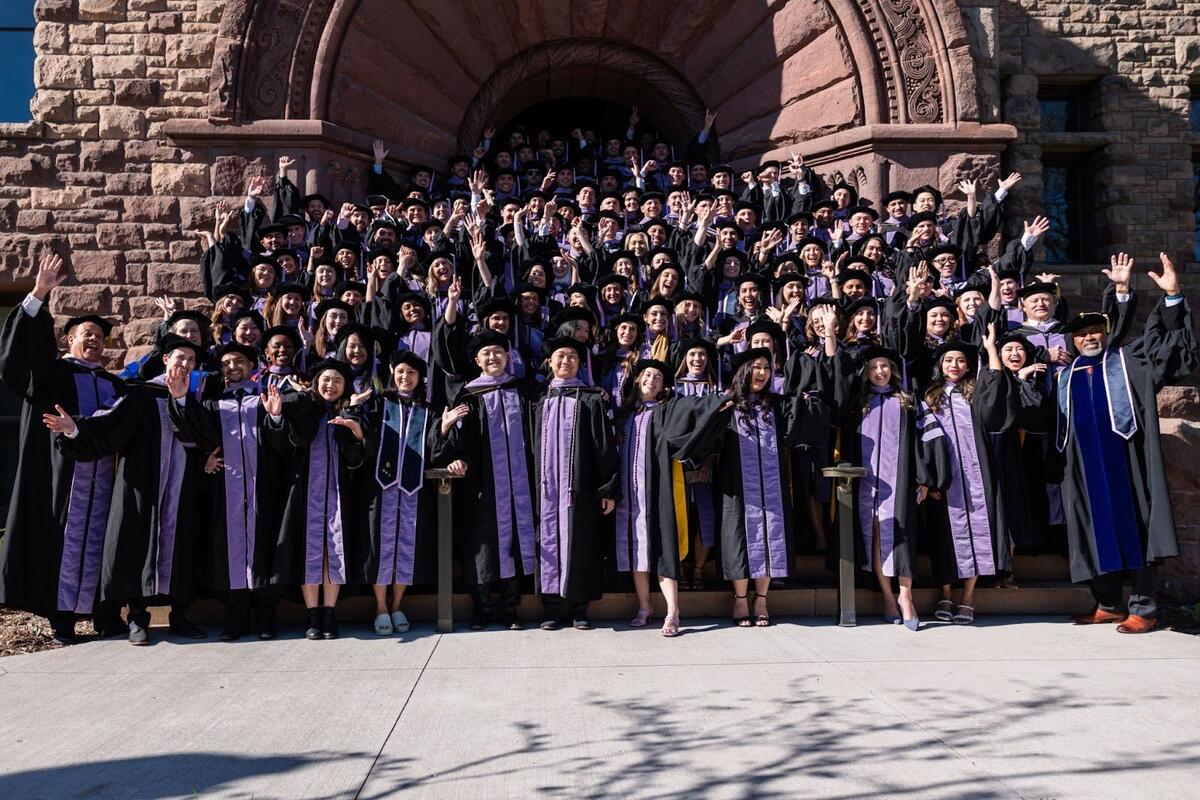 A large group of DDS graduates celebrate on the steps of a brick building in their regalia