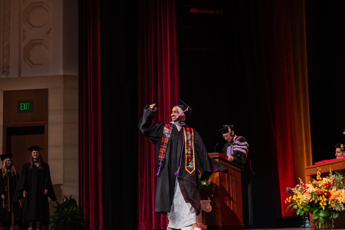 Student waves at the audience as she crosses the stage at commencement