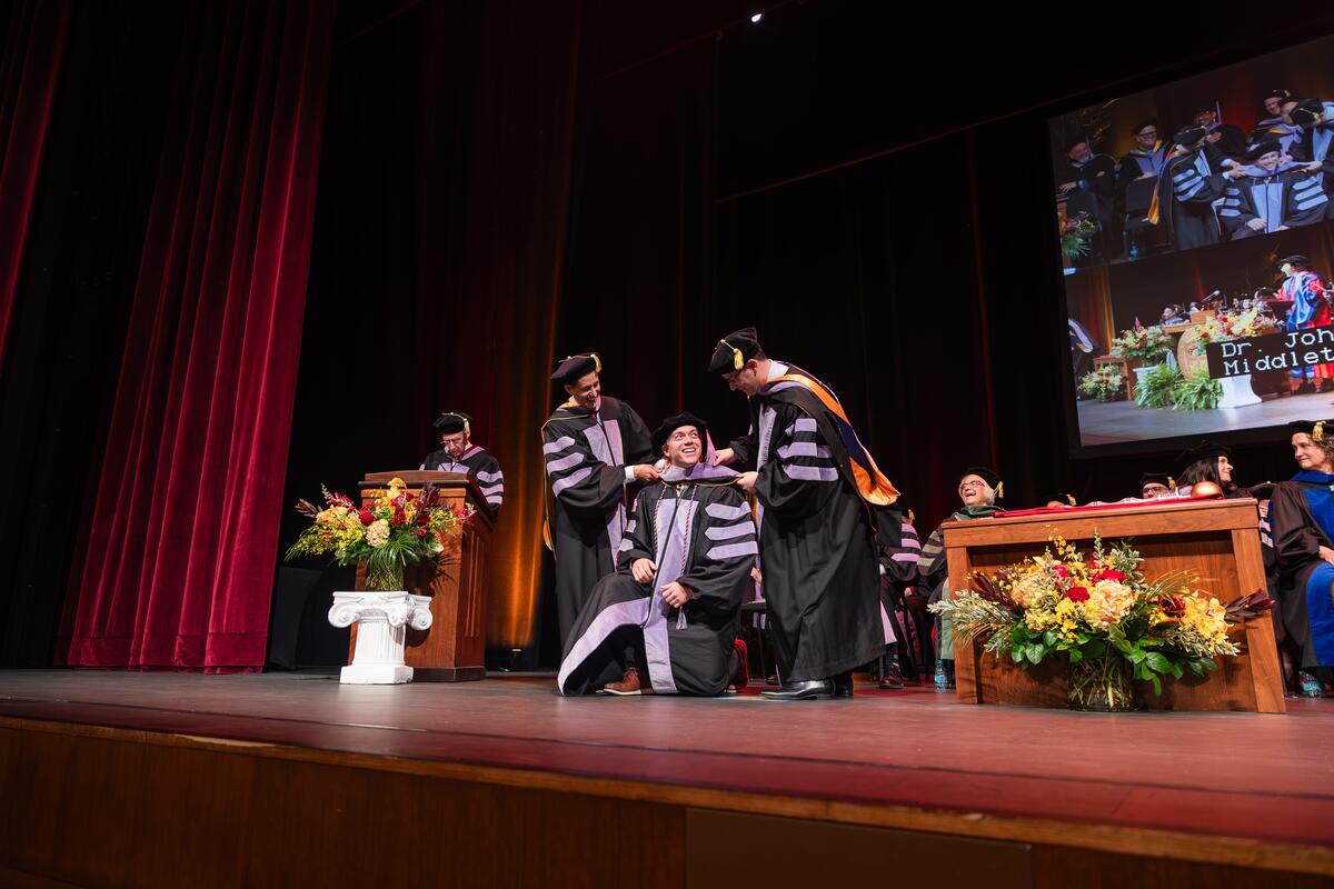 A graduate kneels to receive his doctoral hood