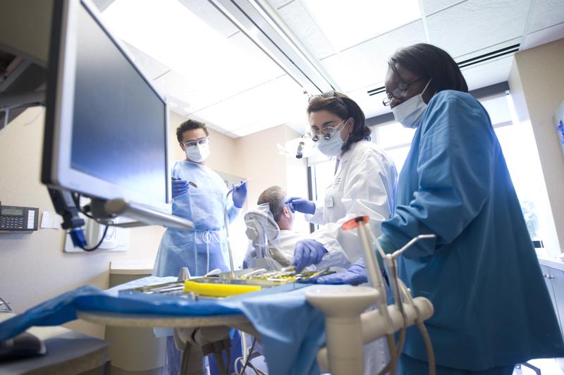 three dentists consult a monitor while patient sits in chair with back to camera