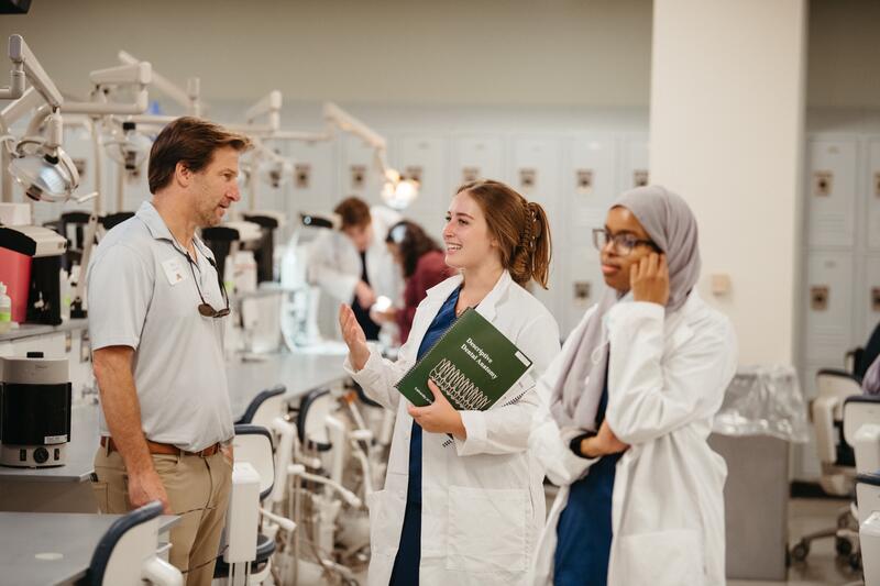 A School of Dentistry alumnus talks to two dental students in white coats while in the clinic