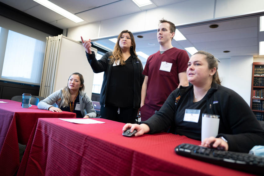 Four residents sit or stand in a conference room and discuss what is displayed on a projector screen