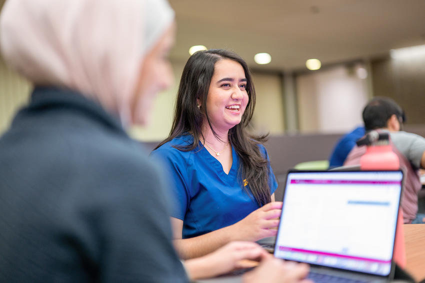 A student smiles and talks with other students while another student works on her computer, blurred in the foreground