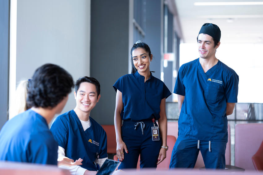 Four students in navy blue scrubs gather and talk around a lounge space in HSEC