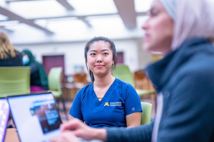 Two students talk at a table (one blurred) with laptops