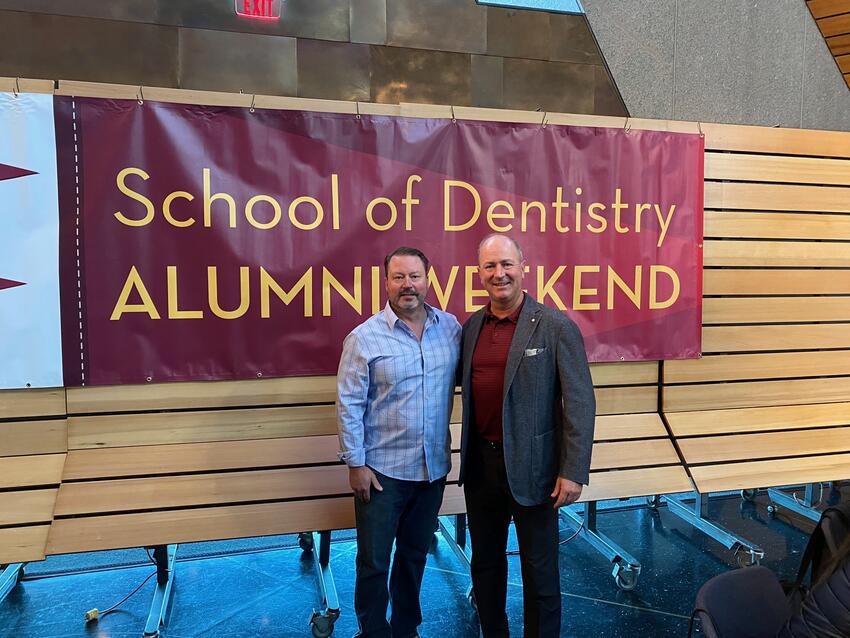 Ryan anderson and Mark Bachman in front of an Alumni Weekend banner